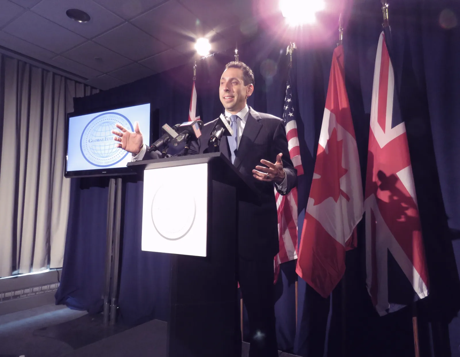 Man speaking at podium with multiple flags and presentation screen visible.