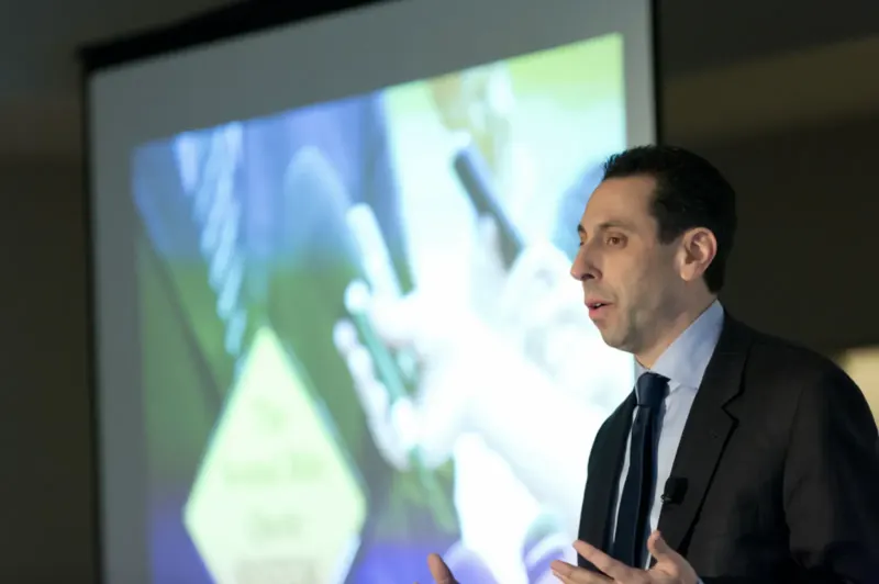 Jess Todtfeld mid-talk, gesturing with both hands, a projected slide behind him